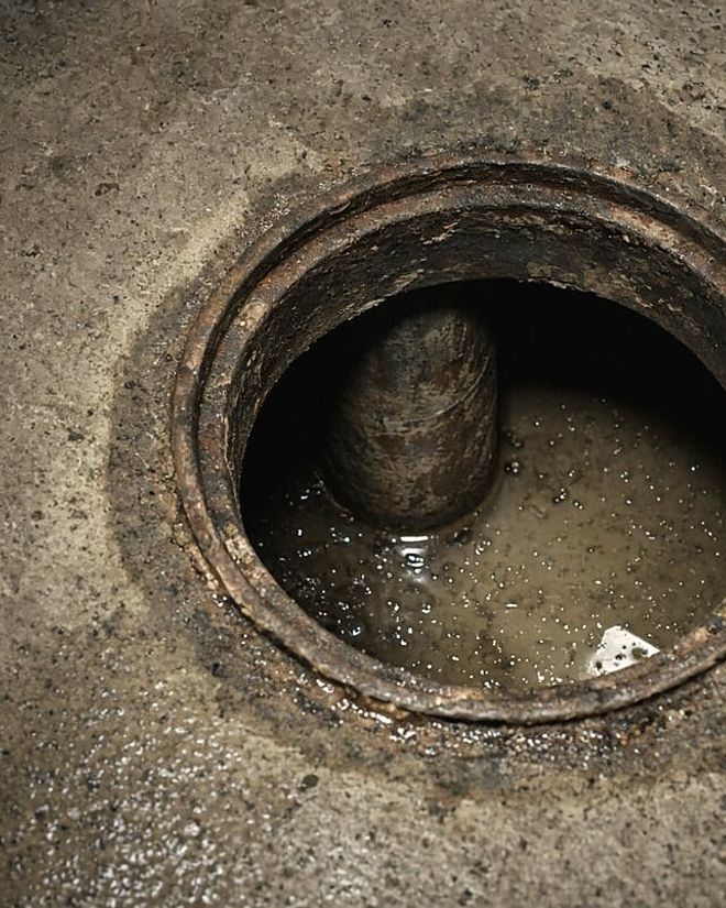 Open rusty sewer cleanout in a concrete basement floor with standing wastewater inside the pipe